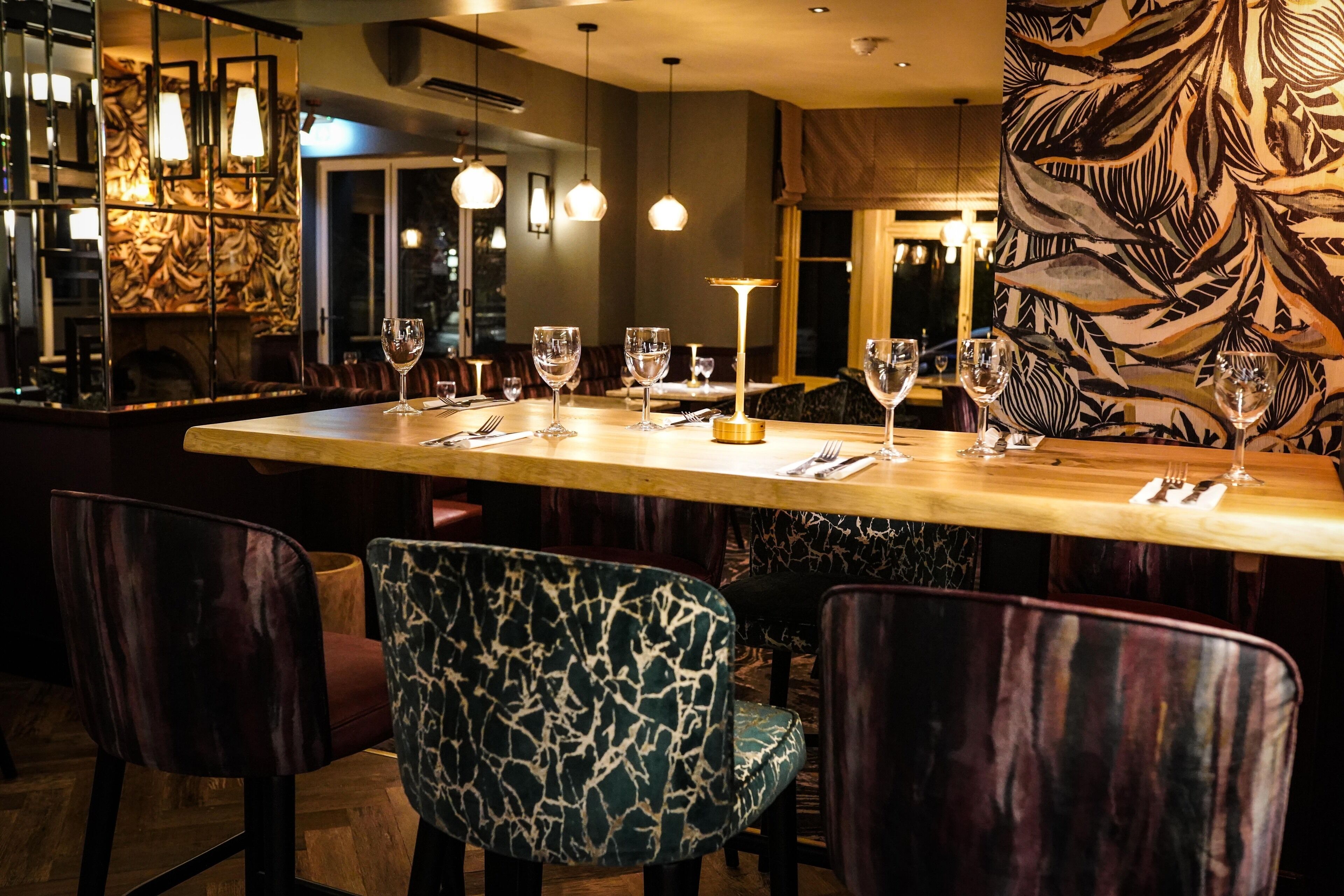 A restaurant with a long wooden table, several chairs, and wine glasses set up.