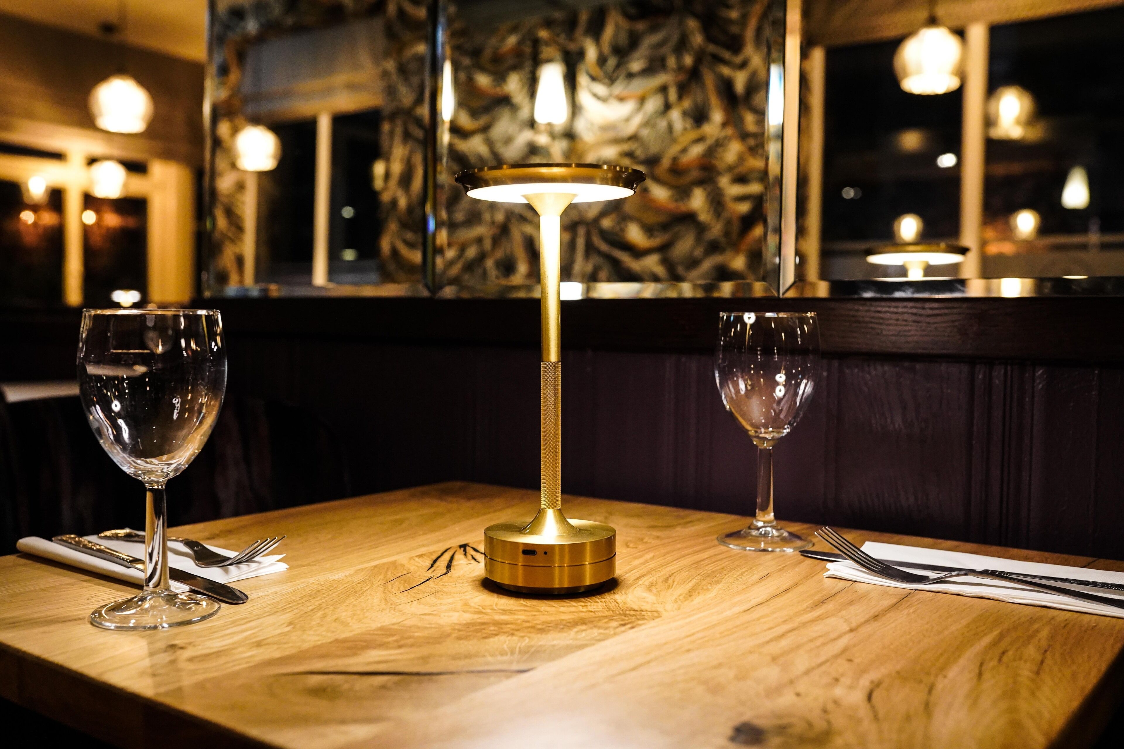 Une table en bois sur laquelle se trouve une lampe de table de couleur dorée, deux verres à vin vides et un ensemble de couverts.