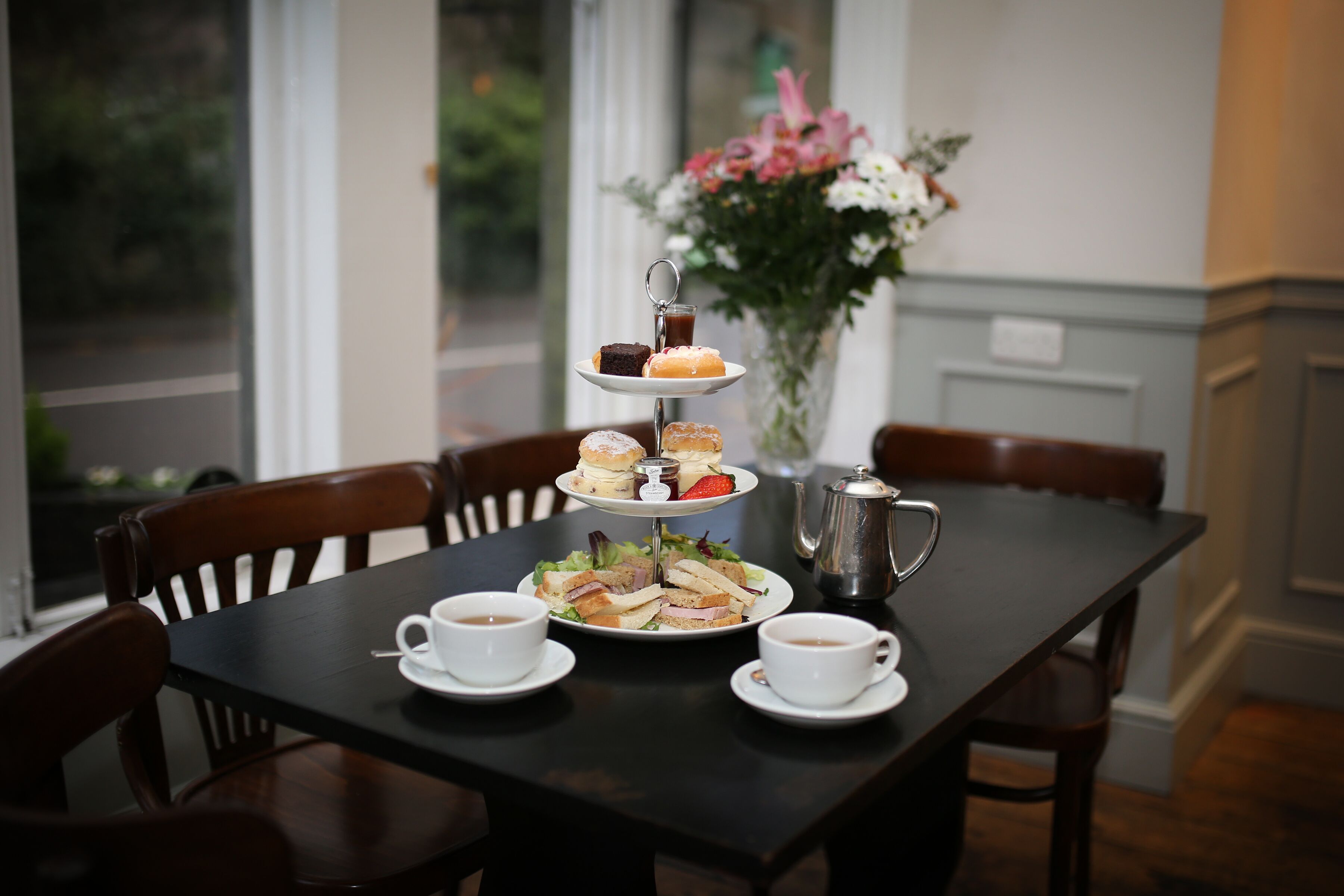 A three-tiered dessert stand with sandwiches and a coffee pot on a table.