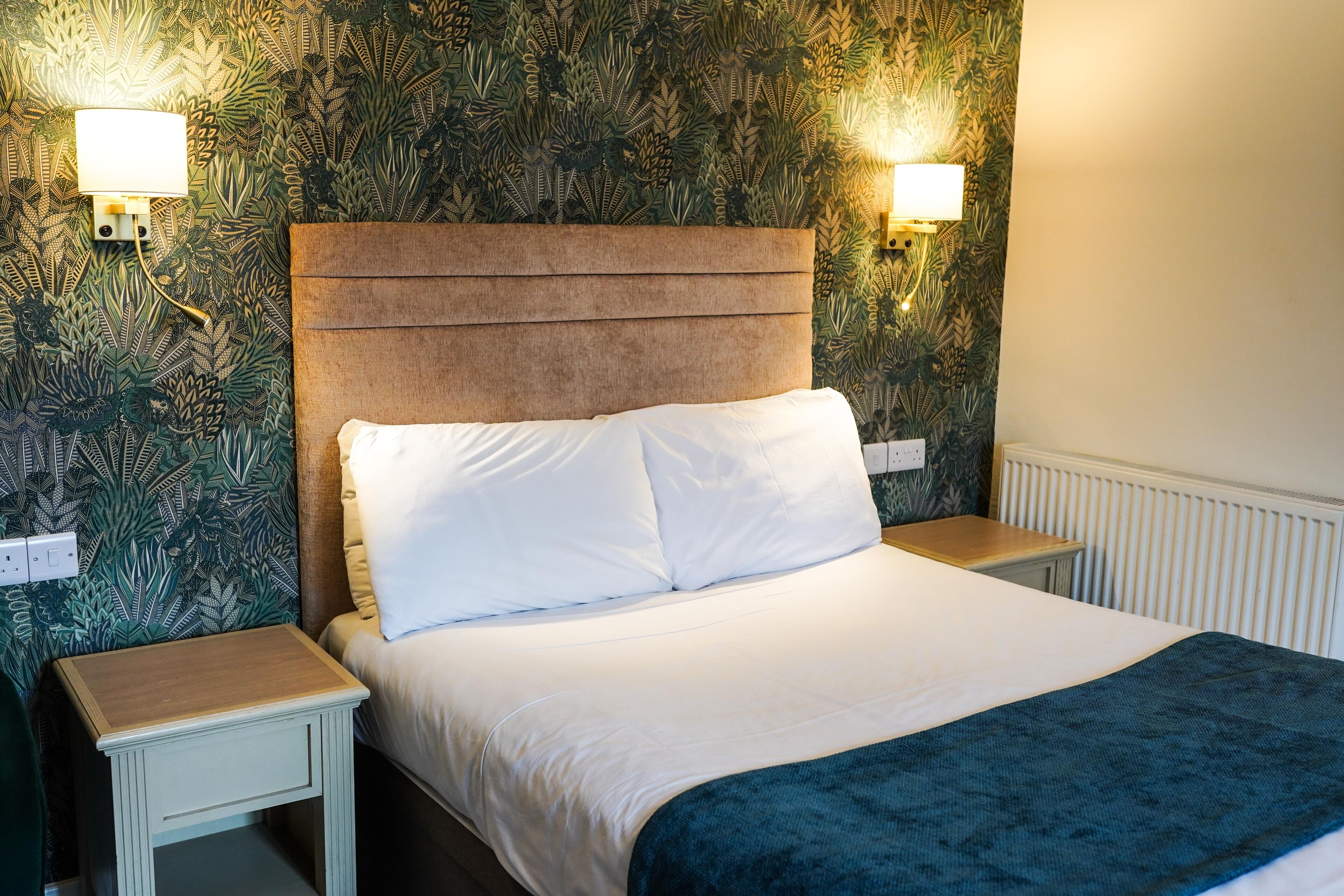 A neatly made bed with white linens and a blue throw blanket, flanked by two bedside tables with lamps, set against a wall with a leafy pattern wallpaper.