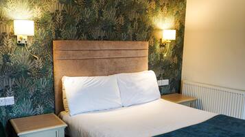 A neatly made bed with white linens and a blue throw blanket, flanked by two bedside tables with lamps, set against a wall with a leafy pattern wallpaper.