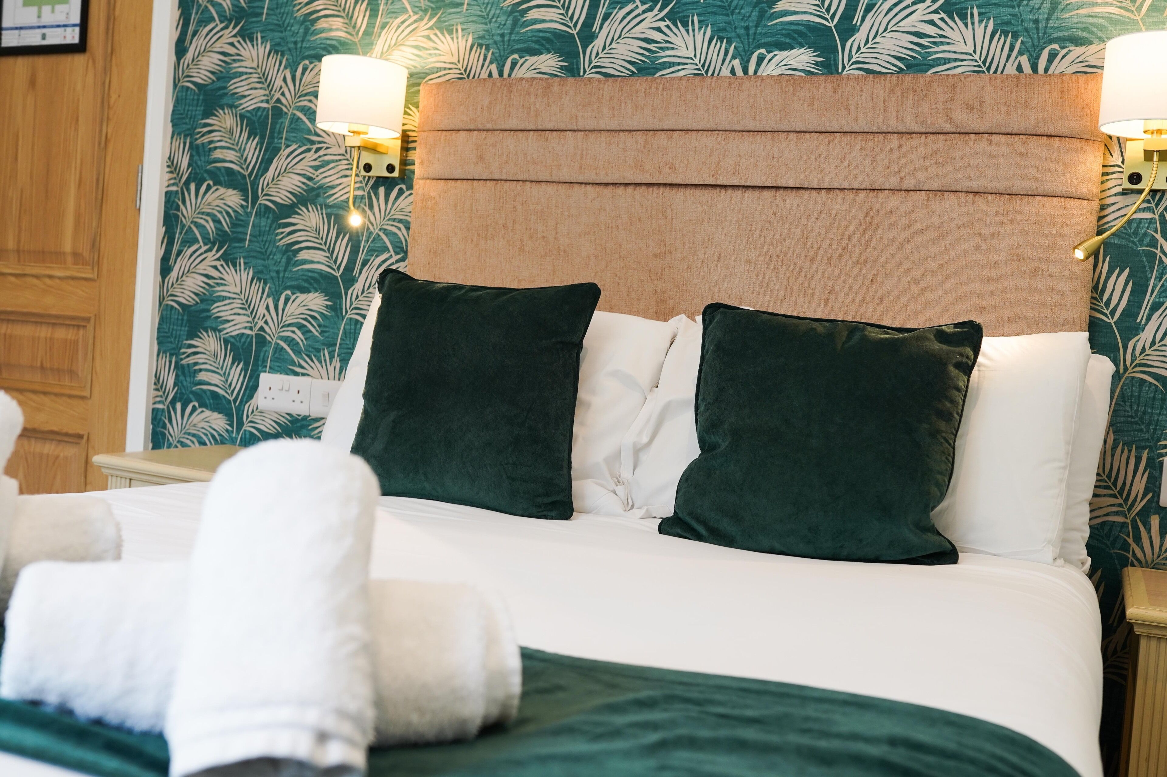 Una cama bien hecha con ropa blanca y almohadas verdes, un cabecero con panel marrón claro y una pared con estampado de hojas.