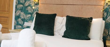 Una cama bien hecha con ropa blanca y almohadas verdes, un cabecero con panel marrón claro y una pared con estampado de hojas.