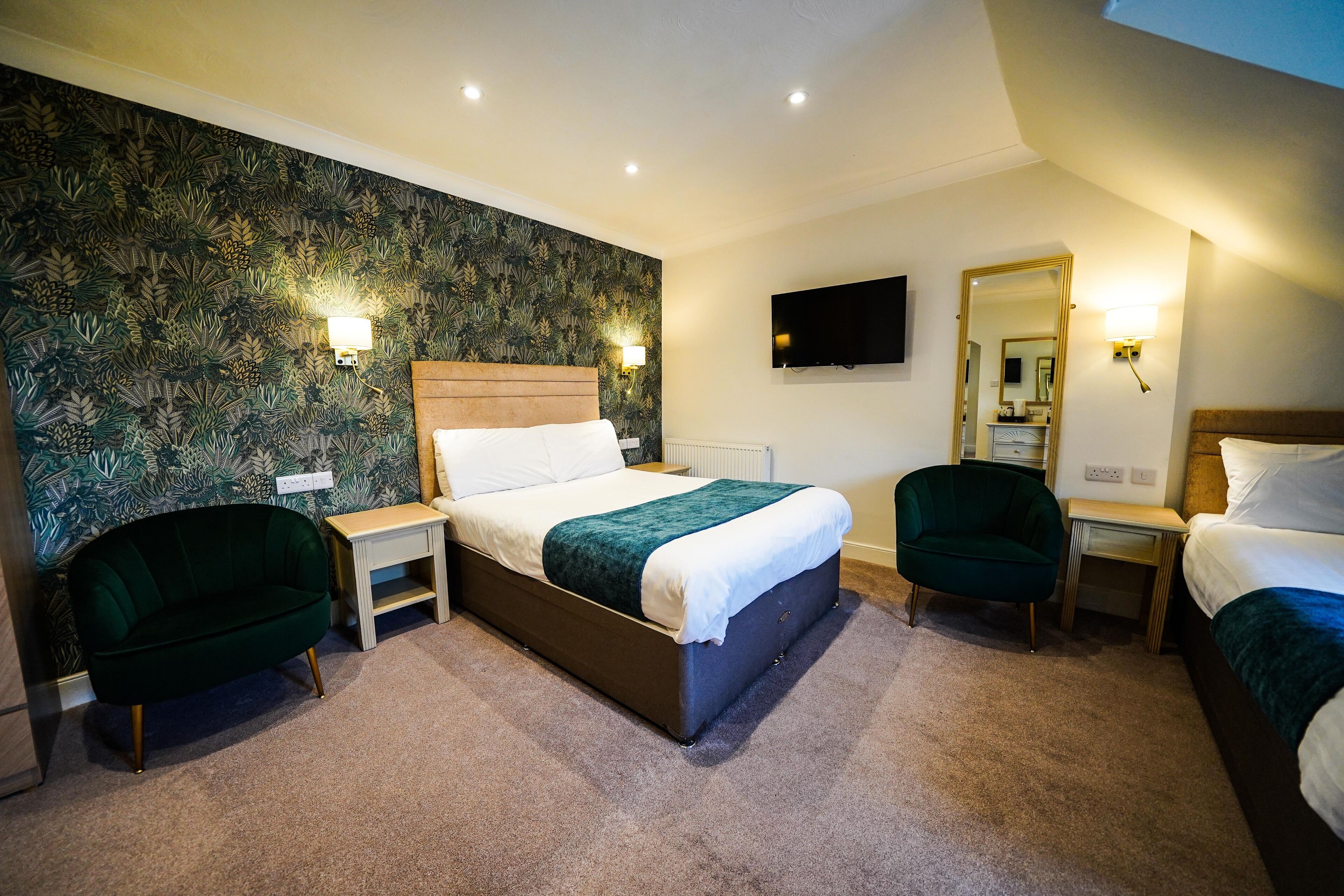 Una habitación de hotel con una cama grande, dos mesitas de noche, un sillón verde, un televisor montado en la pared y un espejo.
