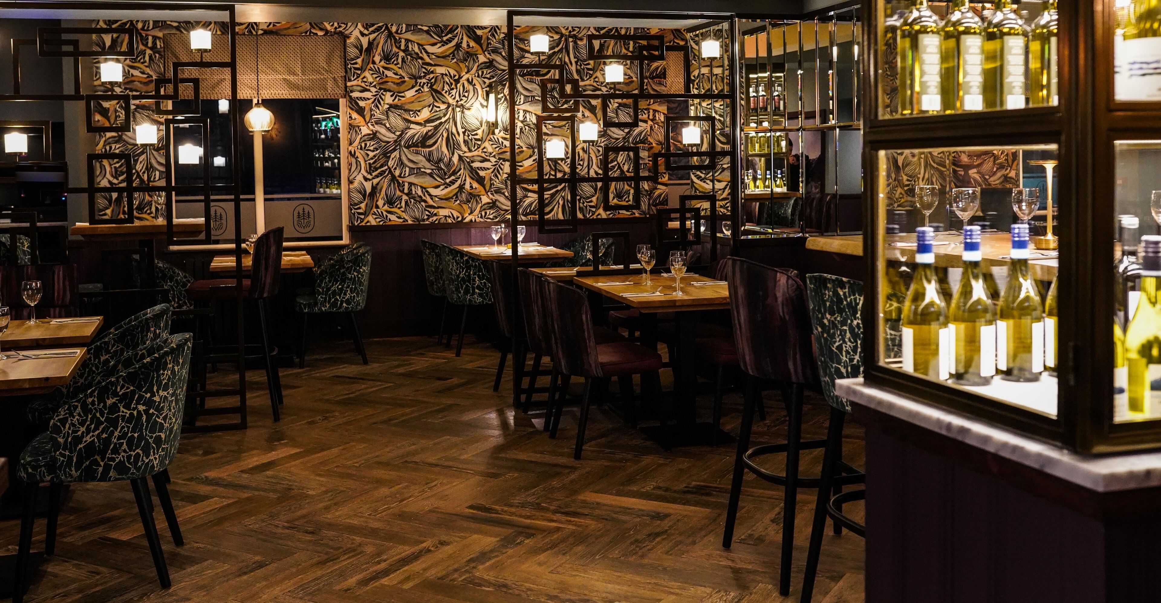 A restaurant interior with wooden flooring, patterned walls, and a bar area with wine bottles.
