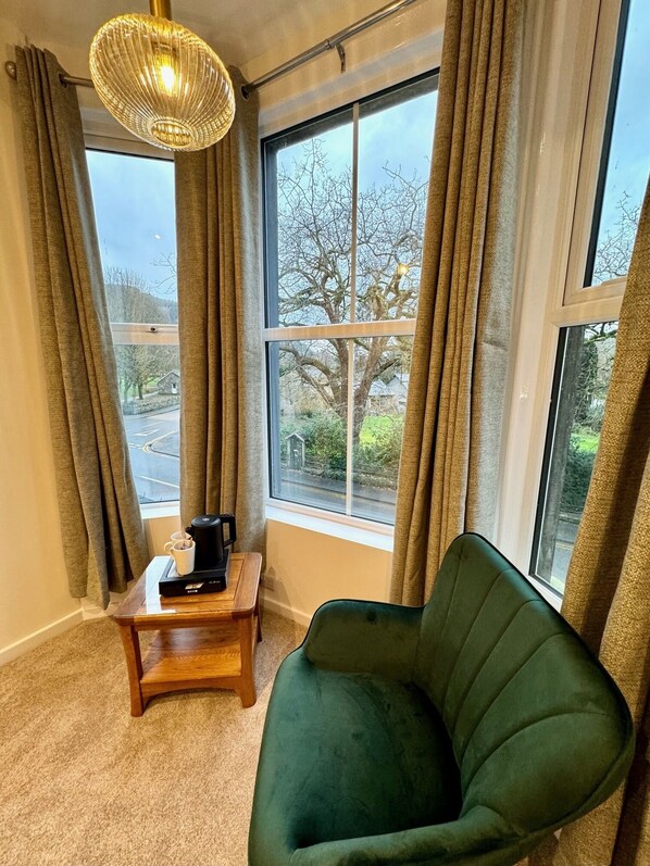 A room with a large window, a green armchair, a wooden side table with a coffee maker, and a hanging light fixture.