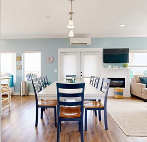 A dining area with a long table and chairs, a clock on the wall, and a television in the background.
