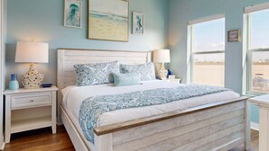 A bedroom with a wooden bed, two bedside tables with lamps, a window with a view, and three wall art pieces. - Views of Ocean, Boardwalk to Beach and Golf Cart Included, Private Heated Pool, Pet Friendly (Port Aransas)