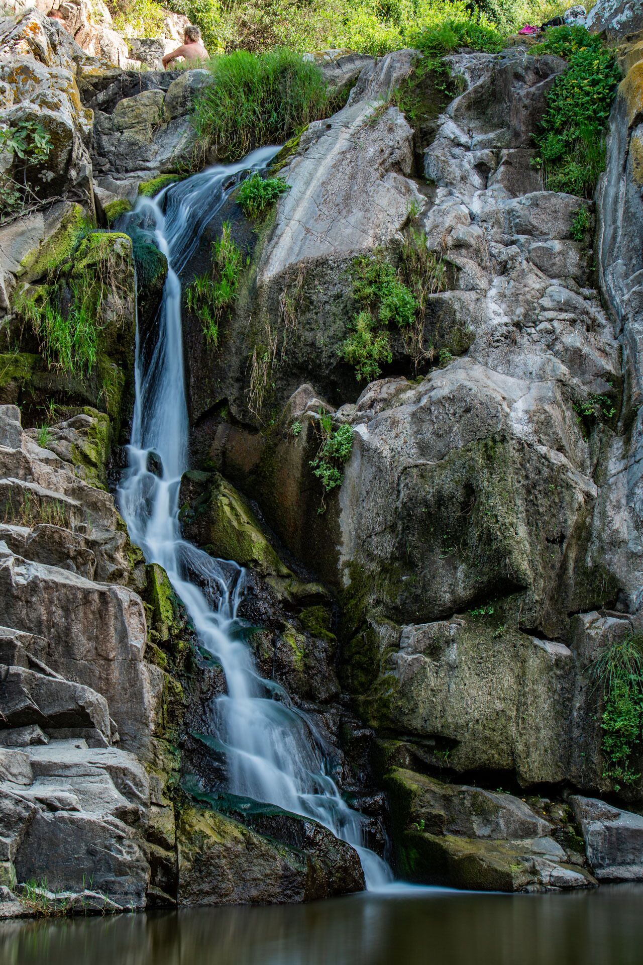 Una cascata che scorre su un terreno roccioso, con pietre ricoperte di muschio e vegetazione rigogliosa.