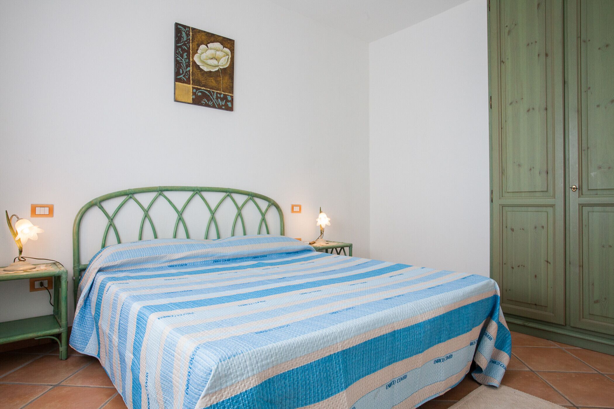 A bedroom with a bed, a nightstand, a lamp, a green wardrobe, and a wall with a floral painting.