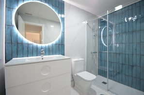 A bathroom with a white vanity, a round mirror, a toilet, and a glass-enclosed shower.