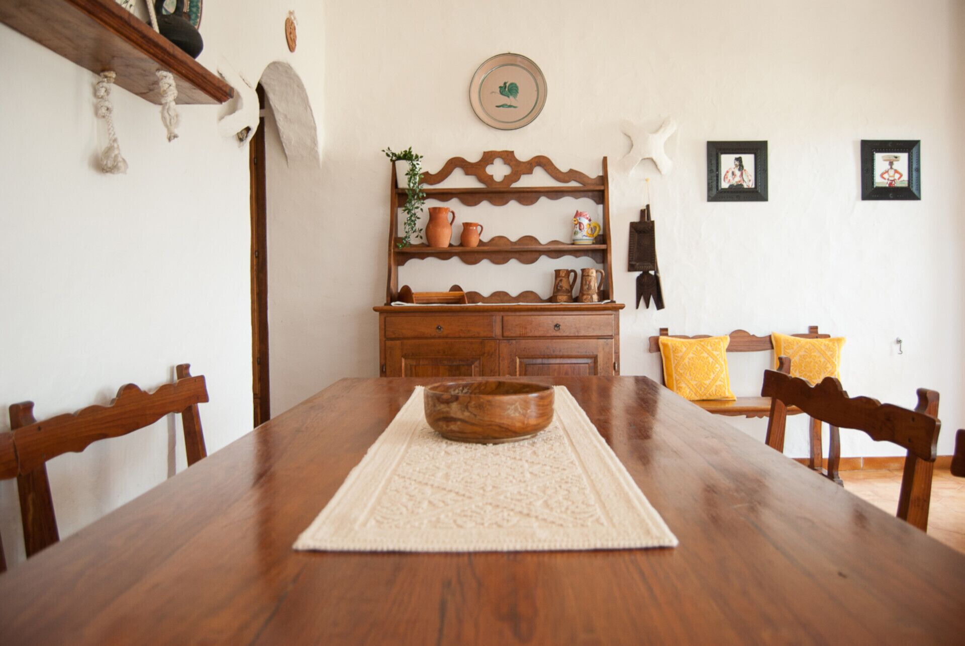 A dining area with a wooden table, chairs, a decorative wooden headboard, a small cabinet with pottery, and a wall with framed pictures.