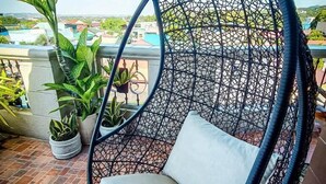 A black wicker swing chair with a cushion on a tiled patio with potted plants and a view of buildings. - Overlooking (Antipolo)