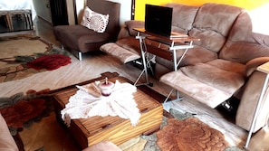 Una sala de estar con un sofá marrón, una mesa de centro de madera con mantel de encaje y un ordenador portátil sobre una mesita.