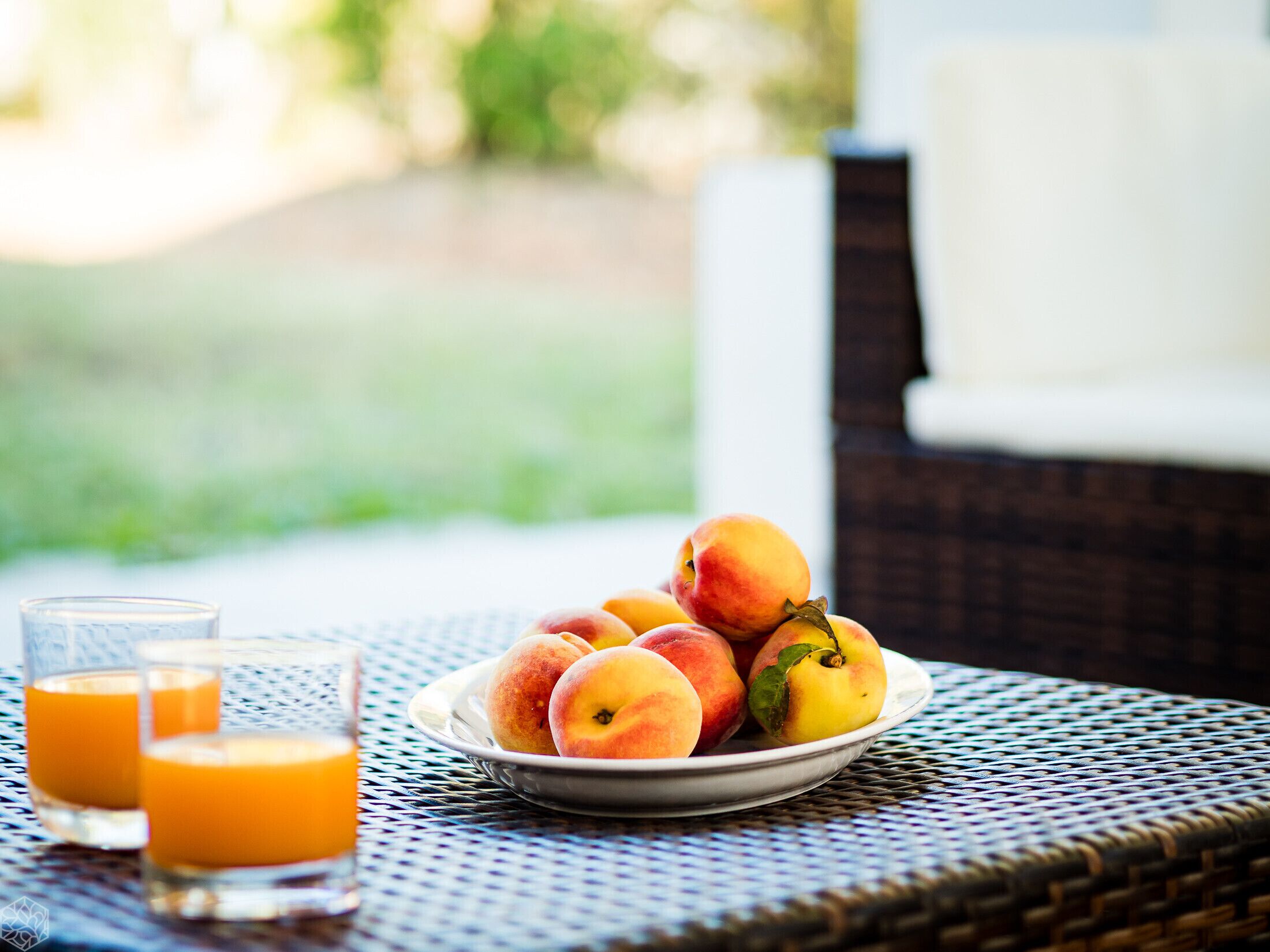 Une table en osier sur laquelle se trouve une assiette de pêches et deux verres de jus d’orange.
