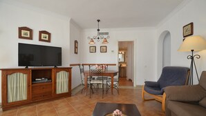 Un salon comprenant un téléviseur, un meuble en bois, une table à manger, des chaises, un canapé, une lampe et un vase à fleurs.