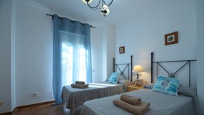 Une chambre avec deux lits, une table de chevet avec une lampe, une fenêtre avec des rideaux et un tableau accroché au mur.