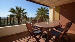 Une table et des chaises en bois sur un balcon donnant sur des palmiers et la mer.