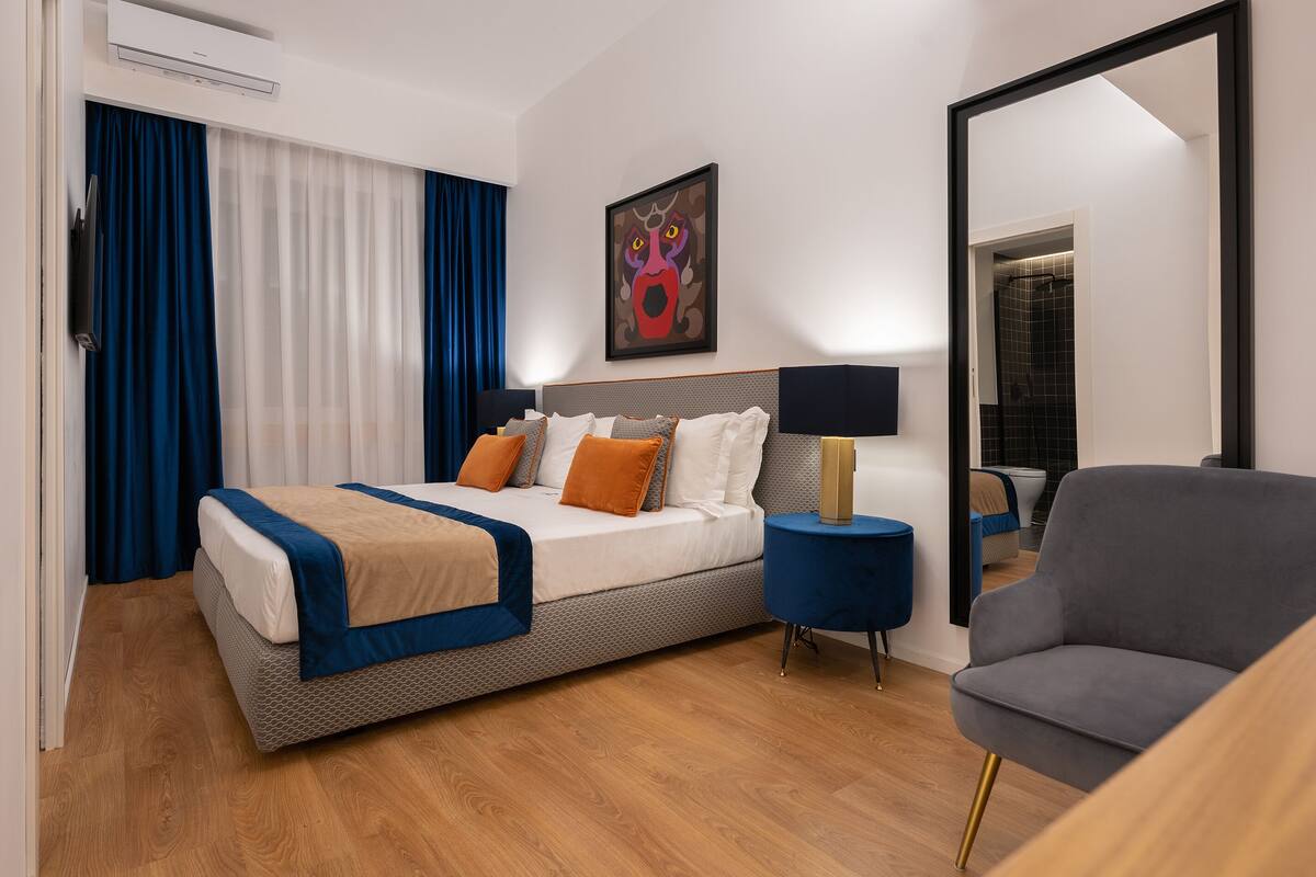 A modern hotel room with a large bed, a side table with a lamp, a chair, and a large mirror.