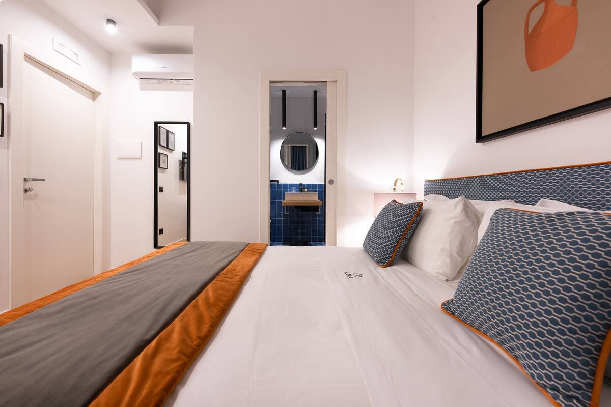 A modern hotel room with a neatly made bed, a bathroom with blue tiles, and a framed picture on the wall.