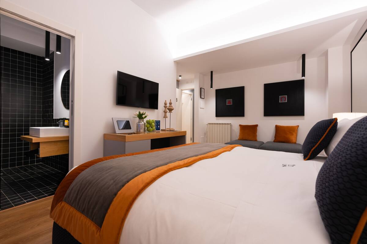 A modern hotel room with a large bed, a bedside table, a flat-screen TV, and a bathroom with a black tiled shower area.