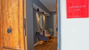 Un interior de hotel moderno con una puerta de madera, una pared gris, un espejo redondo, un sillón gris y una pequeña mesa.