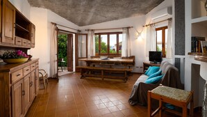 Una stanza accogliente con un tavolo da pranzo in legno, un divano e una TV. Si vede una finestra con tende e vista sul verde circostante.