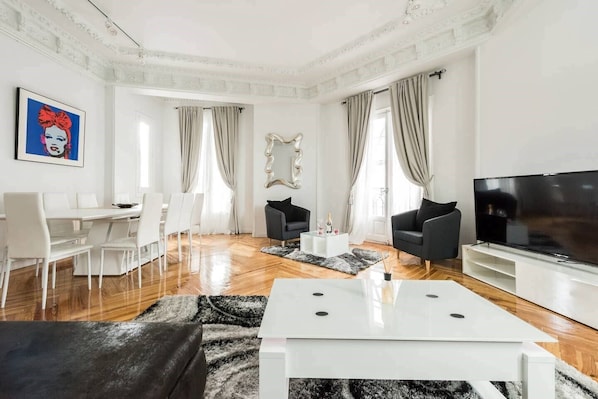 A modern living room with a white TV stand, black sofas, a dining table with chairs, and a large mirror.