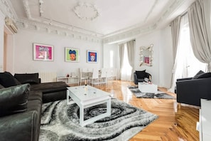 A spacious living room with a modern design, featuring a black leather sofa, a white coffee table, and a black and white rug. There are three framed artworks on the wall, and the room has a large window with curtains.