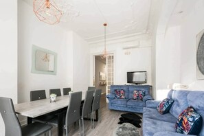 A modern living room with a dining area, a blue sofa with colorful cushions, a flat-screen TV, and a geometric pendant light.