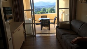 A room with a balcony, a sofa, a table with chairs, and a refrigerator.
