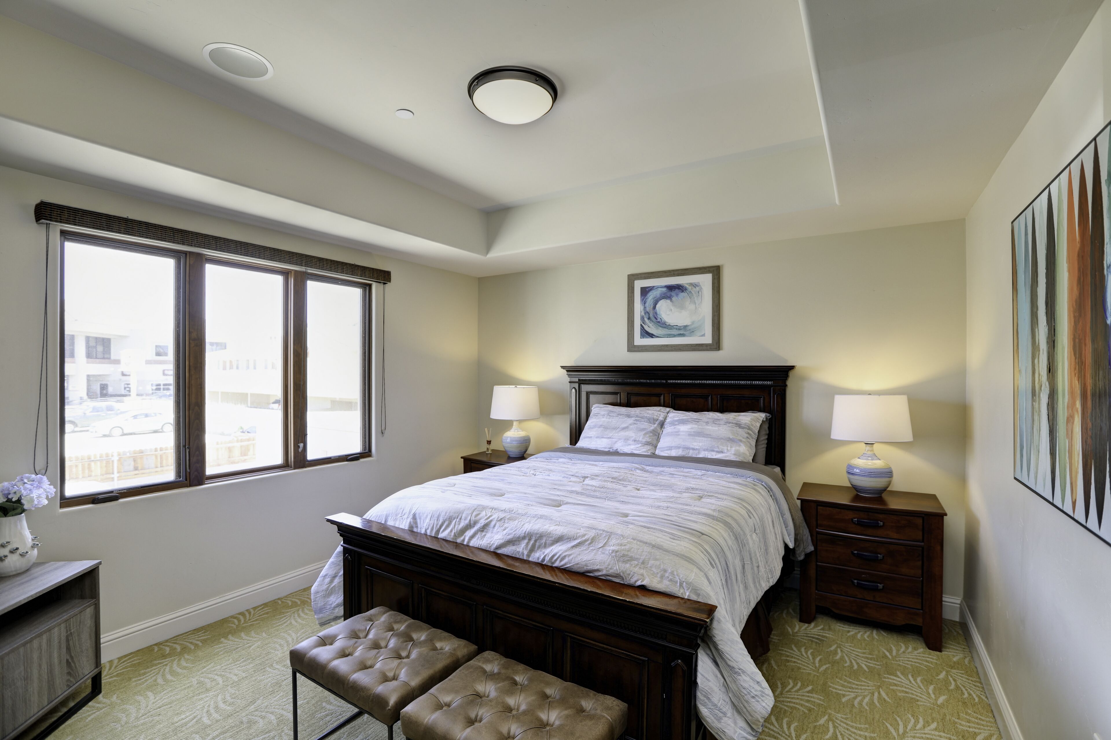 A bedroom with a large bed, two bedside tables with lamps, a window with a view, a framed artwork on the wall, and a bench.