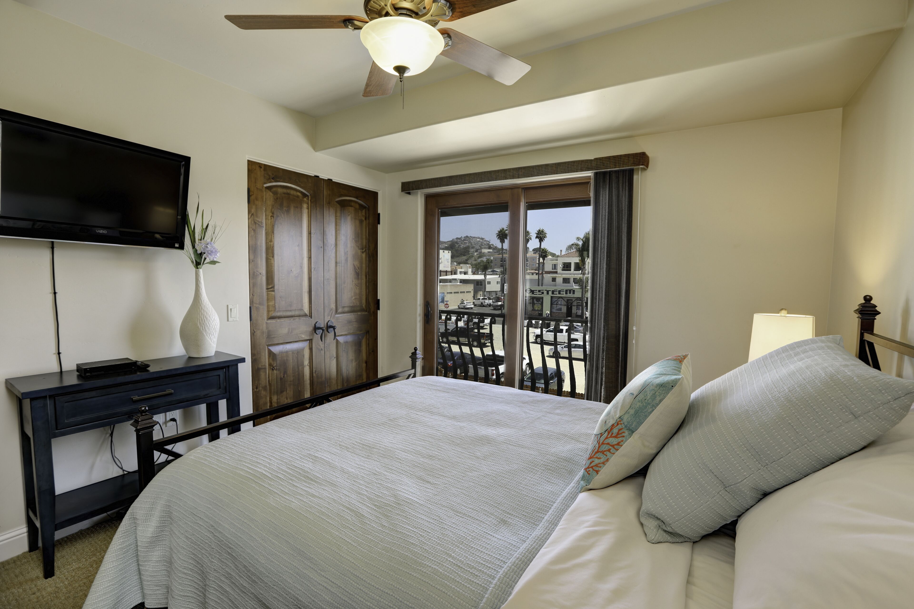 A hotel room with a bed, a desk, a television, a ceiling fan, and a balcony with a view of buildings and palm trees.