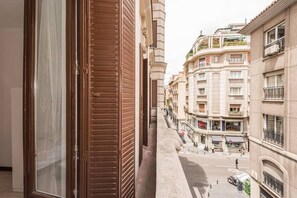 A narrow street view from a balcony with a wooden shutter, overlooking a row of multi-story buildings with balconies and ground-level shops.