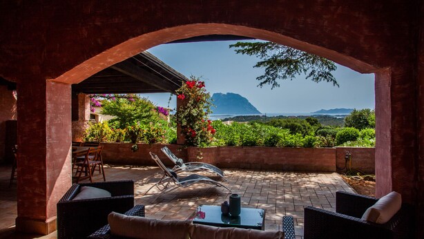 A patio with wicker furniture, a table, and a view of the sea and mountains.