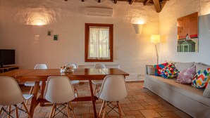 A cozy room with a wooden dining table, white chairs, a sofa with colorful cushions, a TV, and a window with curtains.
