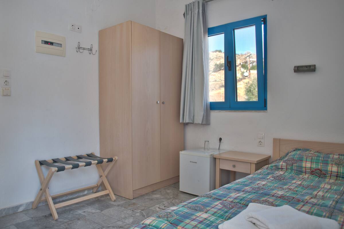 A bedroom with a bed, a desk, a wardrobe, a small fridge, and a window with curtains.