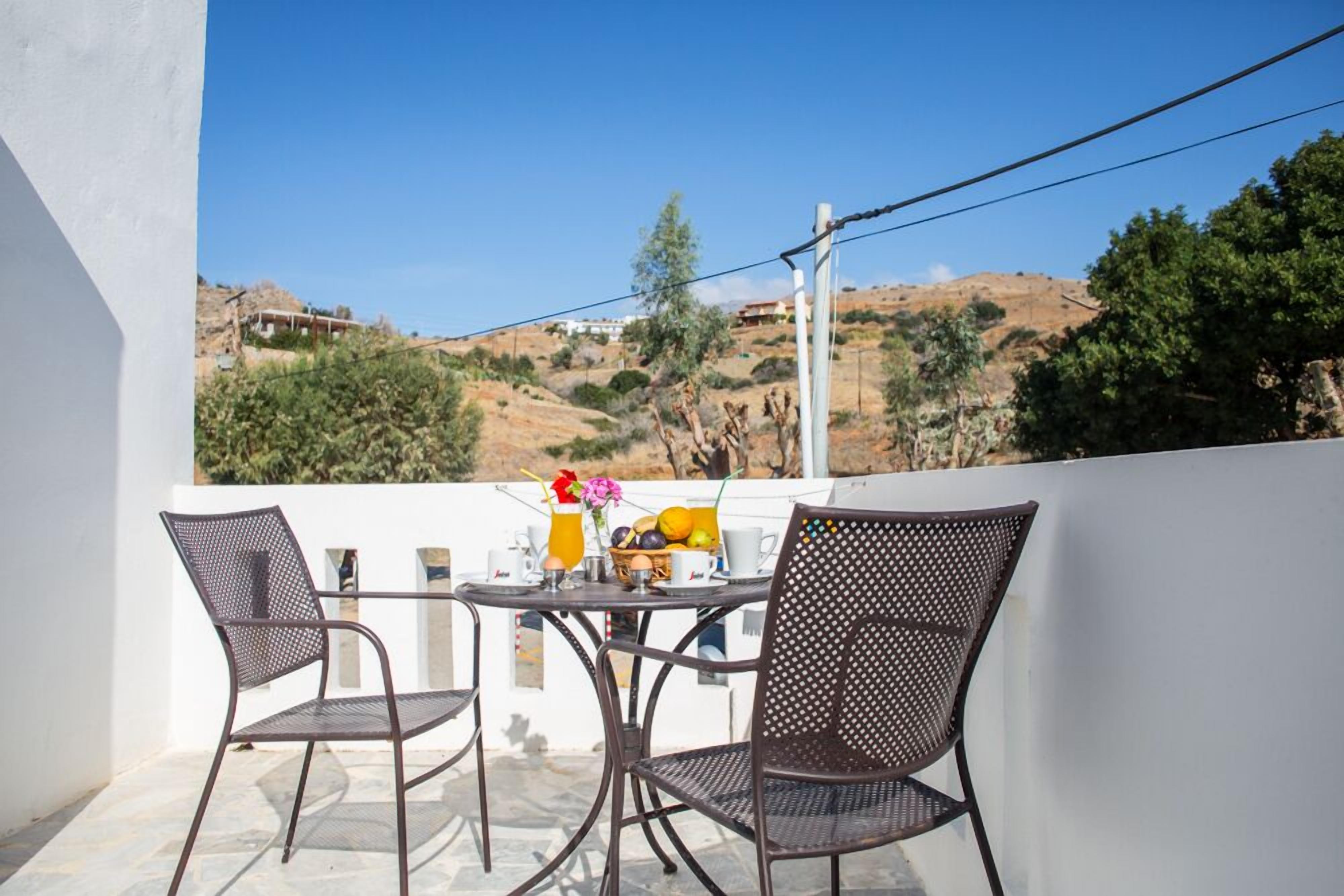 A balcony with two chairs and a table, overlooking a hilly landscape with sparse vegetation and a few buildings in the distance.