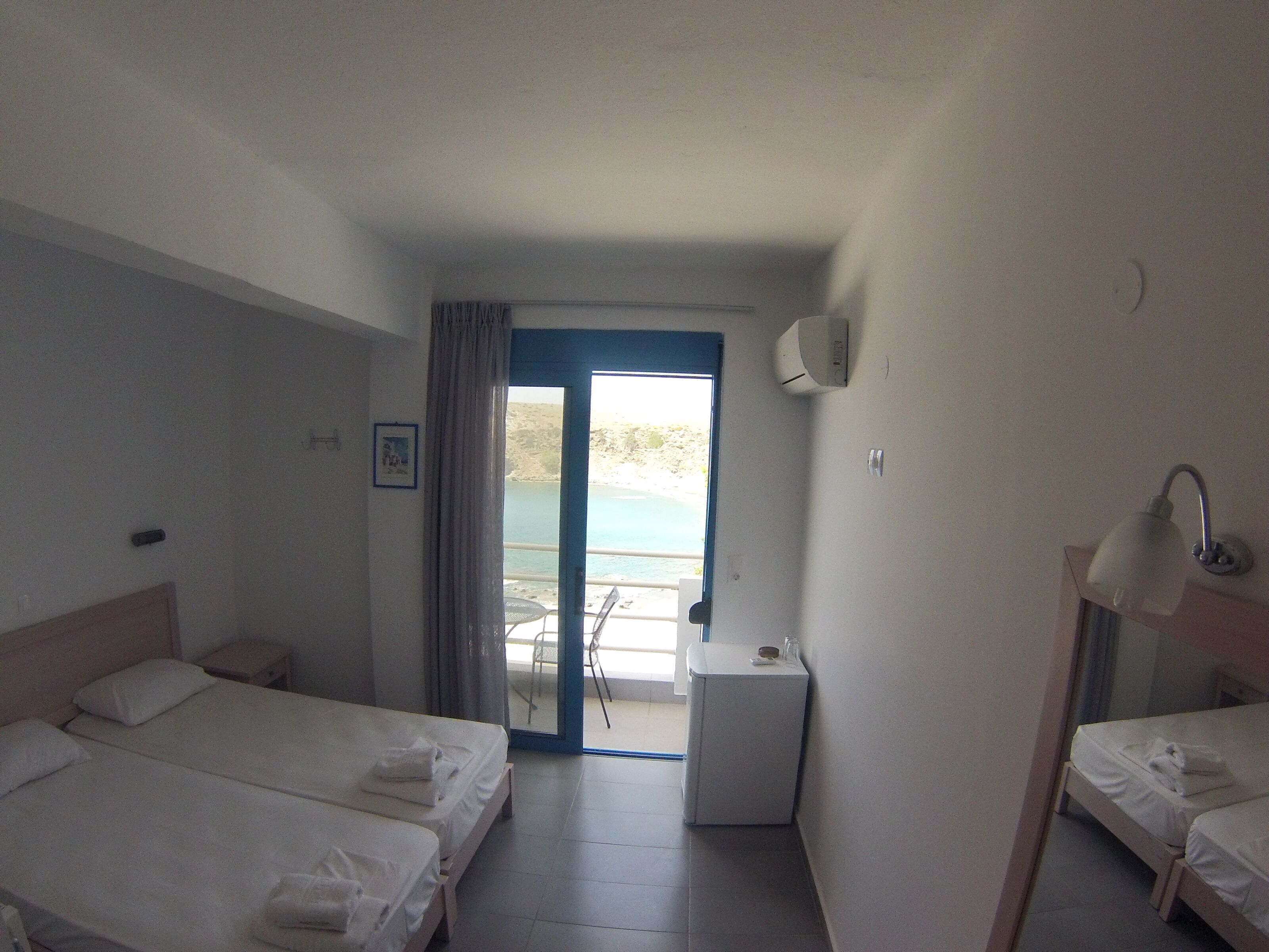 Camera d'albergo con due letti, un tavolino, una sedia, un condizionatore d'aria e vista sul mare tramite una porta in vetro.