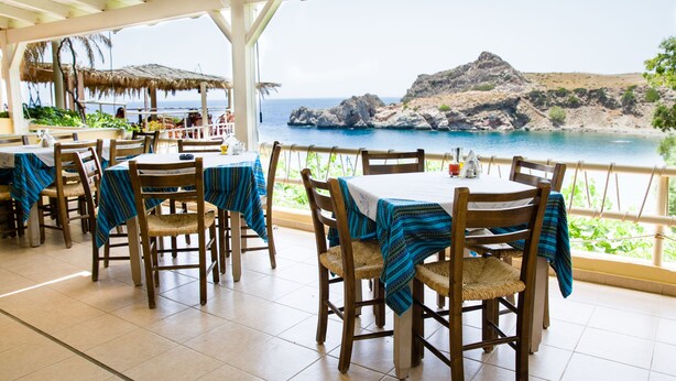 A restaurant with outdoor seating overlooking a coastal view. Tables are covered with blue tablecloths, and wooden chairs are arranged around them. - Agios Pavlos Hotel (Agios Vasileios)
