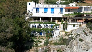 A white building with blue doors and windows, surrounded by trees and rocky terrain. - Agios Pavlos Hotel (Agios Vasileios)