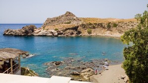 A coastal area with rocky terrain, clear blue waters, and a small beach with a few people. - Agios Pavlos Hotel (Agios Vasileios)