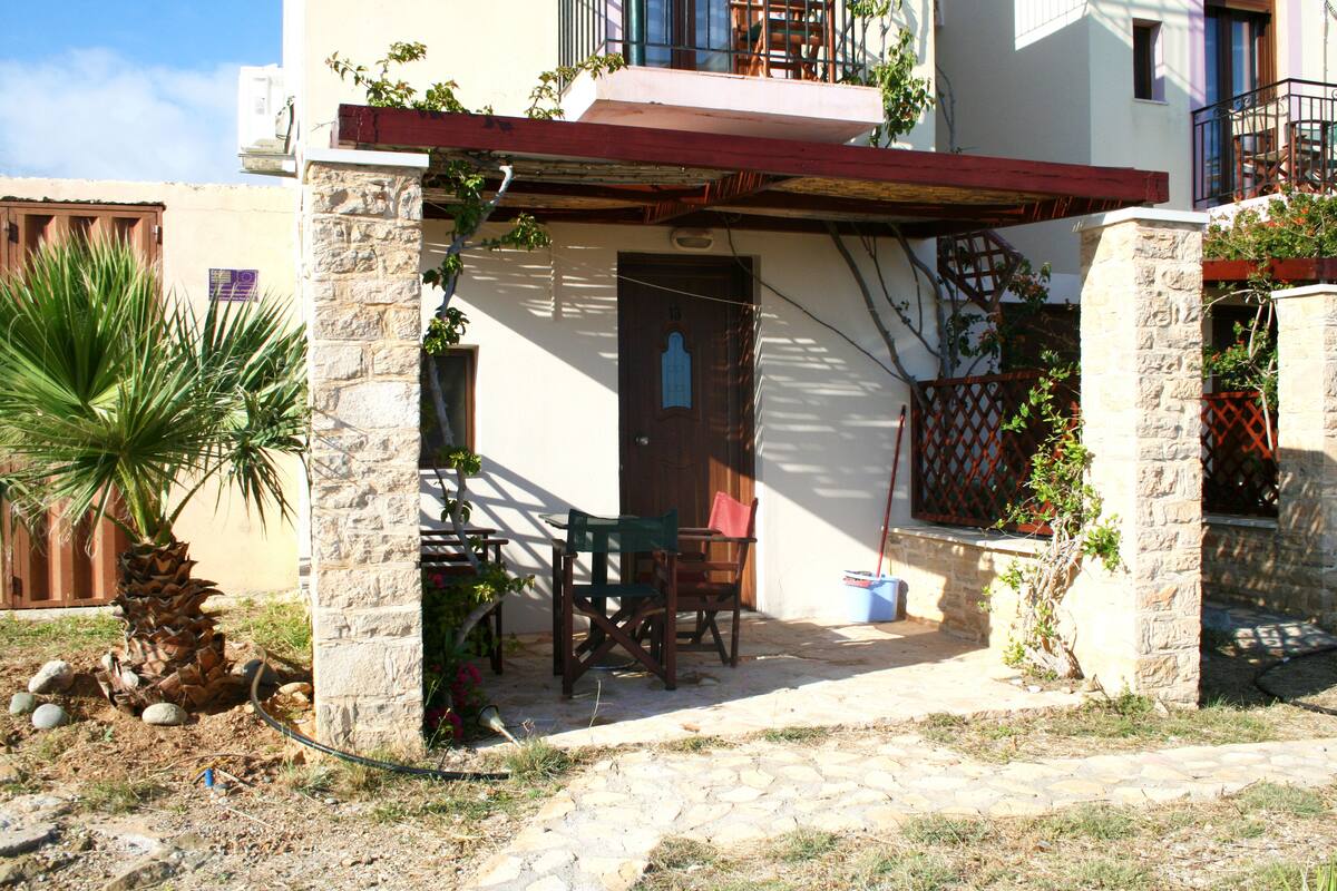 A stone building with a covered patio, a palm tree, and a wooden table and chairs.