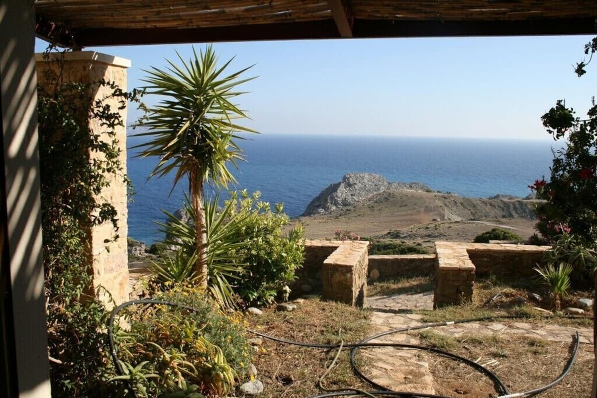 A view from a covered outdoor area with a stone wall, a palm tree, and a view of the sea and a rocky hill.