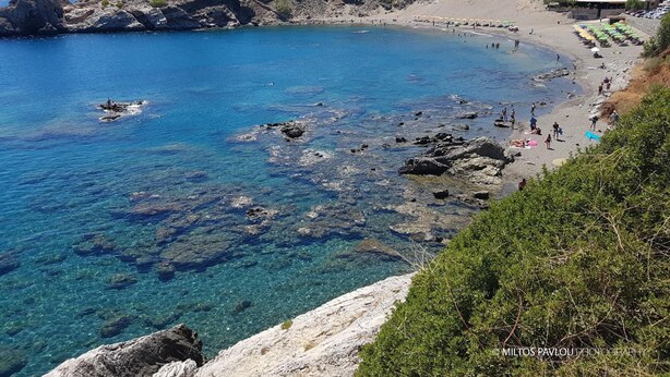 Una playa costera con agua azul cristalina, terreno rocoso y algunas personas disfrutando del lugar.