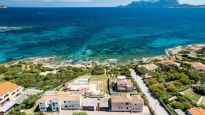 Vista aérea de uma área residencial costeira, com águas azul-claras, linha costeira rochosa e casas com telhados de terracota.