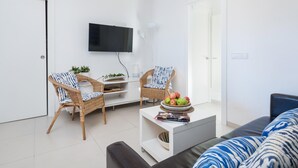 A modern living room with a flat-screen TV mounted on the wall, two wicker chairs with blue and white patterned cushions, a coffee table with a bowl of fruit and magazines, and a sofa with blue and white patterned cushions.