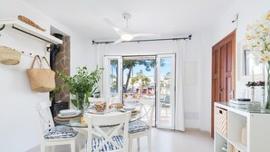 A bright, modern kitchen with white cabinetry, a dining area with white chairs, a ceiling fan, and a view through a glass door to the outside.
