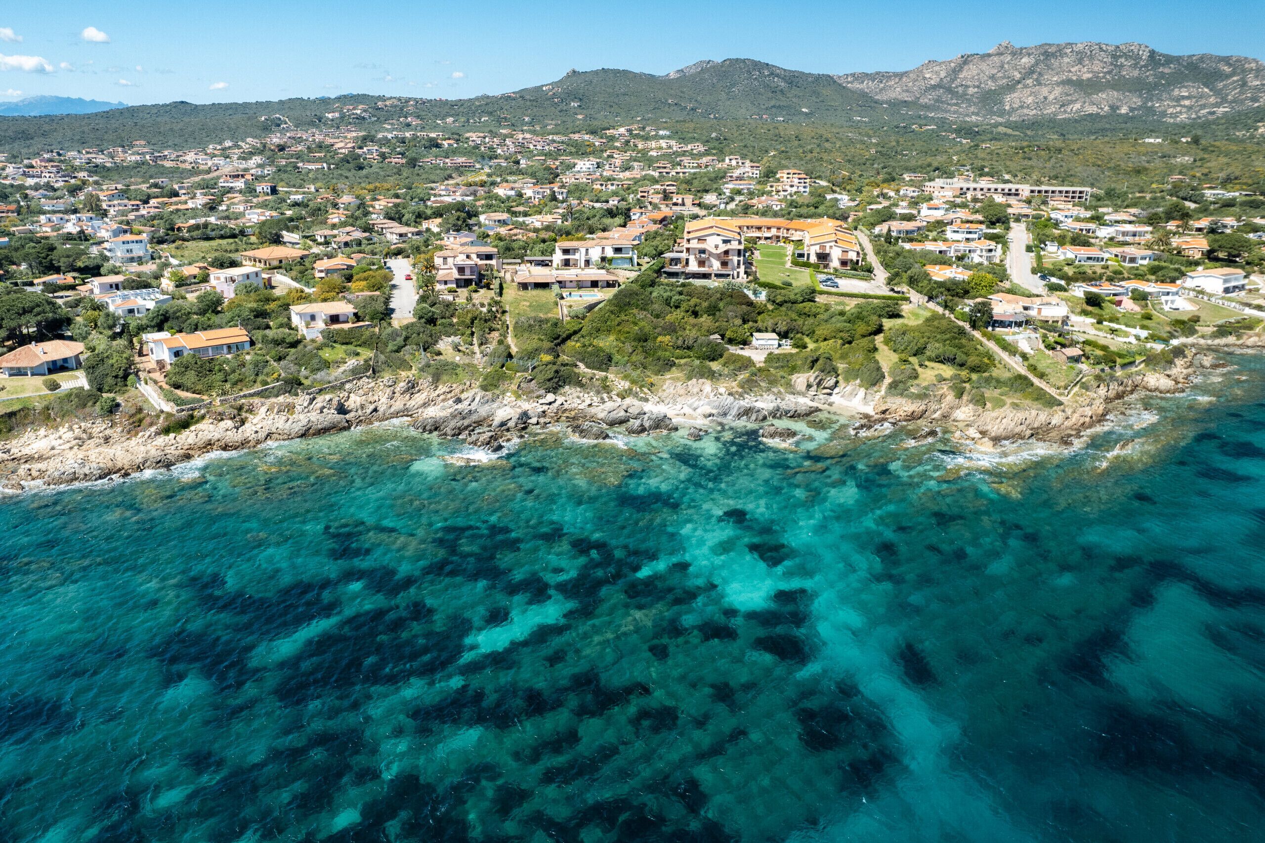Vista aérea de uma área residencial costeira, com casas, águas turquesas translúcidas e colinas verdes ao fundo.
