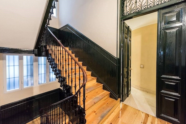 A wooden staircase with black railings leading to a room with a black door and a window with blinds. - TET (Madrid)